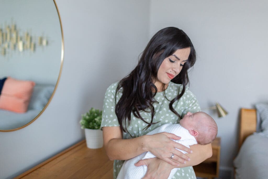 Mother standing in her master bedroom beside a dresser, cradling her newborn and looking down at him during an in-home newborn session in South Jordan, Utah