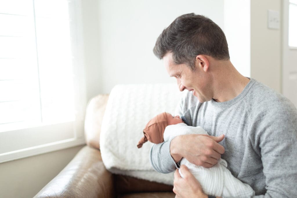 Father sitting in a living room chair, cradling his newborn son and smiling at him during an in-home newborn session in Highland, Utah
