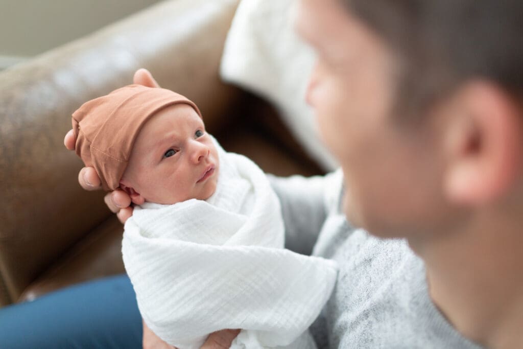 Over-the-shoulder view of a newborn being held in his father’s hands during an in-home lifestyle newborn session in Highland, Utah
