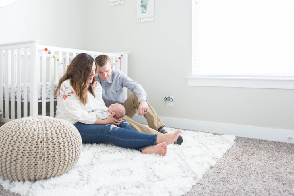 Mom and dad sitting on the nursery floor against the crib, holding and admiring their newborn during an in-home lifestyle session in South Jordan, Utah