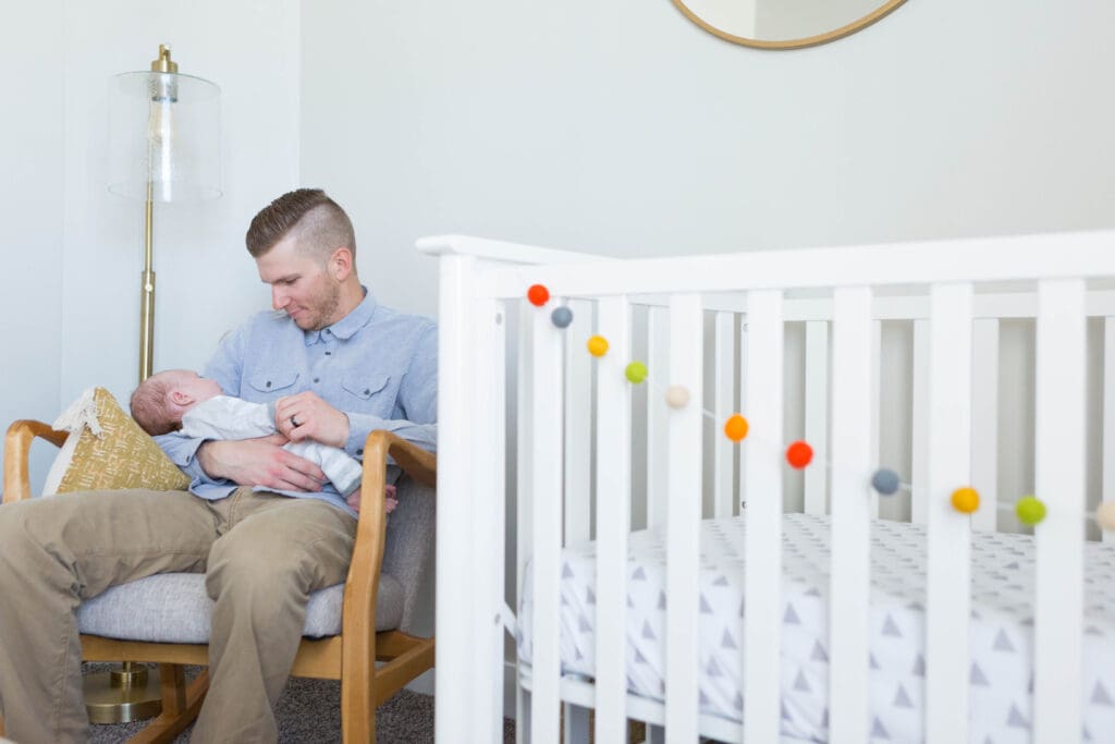 Dad sitting in a nursery rocking chair, holding his newborn during an in-home lifestyle photography session in South Jordan, Utah