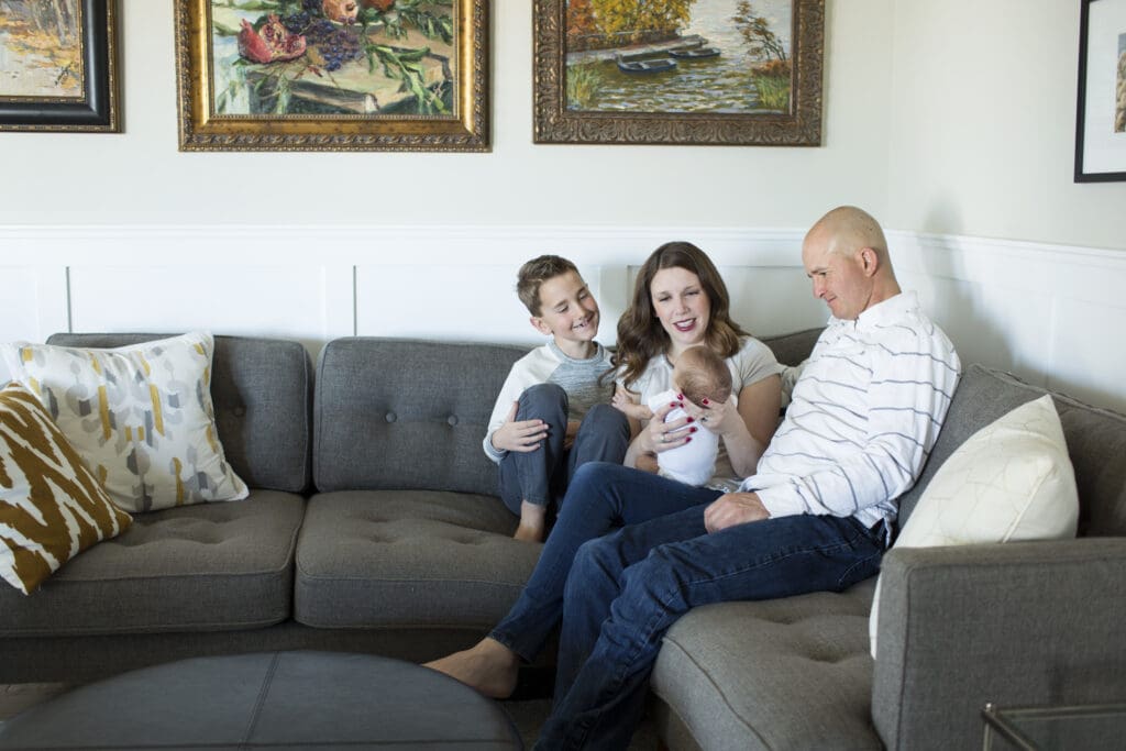 Family sitting on their living room sofa in South Jordan, smiling down at their newborn as mom holds the baby during an in-home lifestyle newborn session.