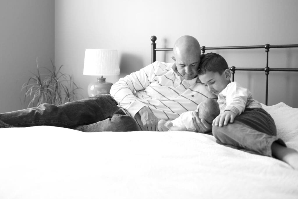 Dad and big brother laying on the master bed, admiring the newborn baby in dad’s hands during an in-home lifestyle session with a Salt Lake newborn photographer.