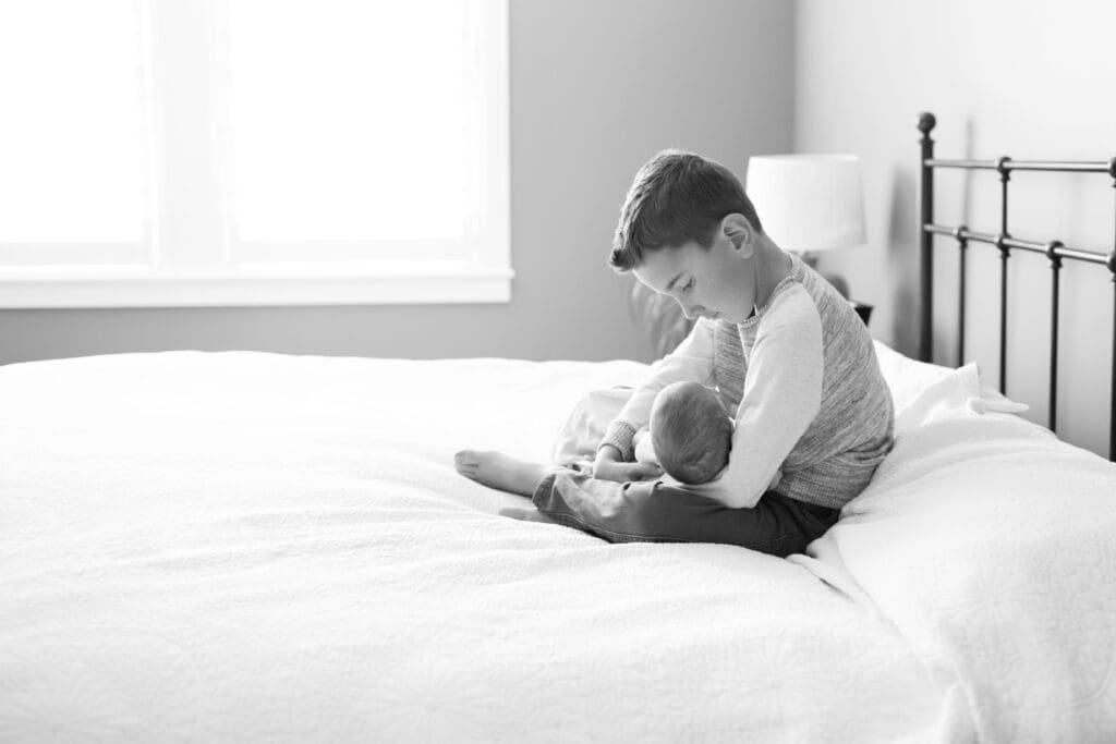 Big brother holding his newborn baby brother on the master bed during an in-home lifestyle session with a Salt Lake newborn photographer.