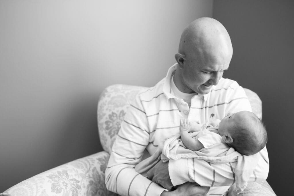 Dad cradling his newborn and gazing into their face while sitting in the rocking chair of the master bedroom during an in-home newborn session in South Jordan, Utah.