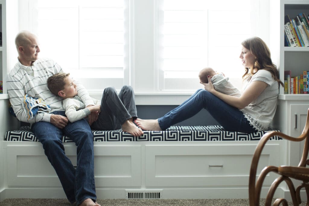 Mom holding newborn while dad and big brother relax beside her on the window seat during a cozy in-home newborn session in their South Jordan nursery.