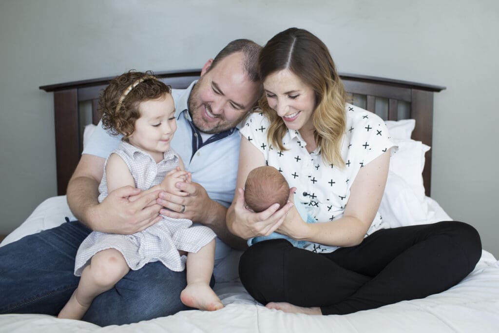 Mom, dad, and toddler sister sitting on a master bed, smiling at their newborn during an in-home newborn photography session