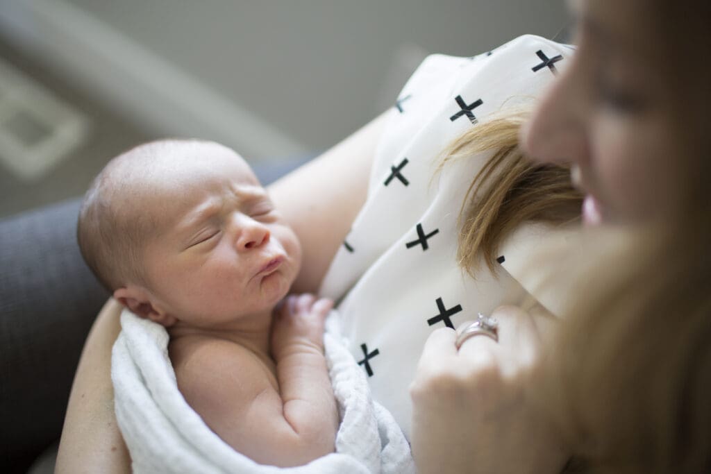 Mother sitting in a rocking chair, cradling her newborn and looking down at his puckered face during an in-home newborn session