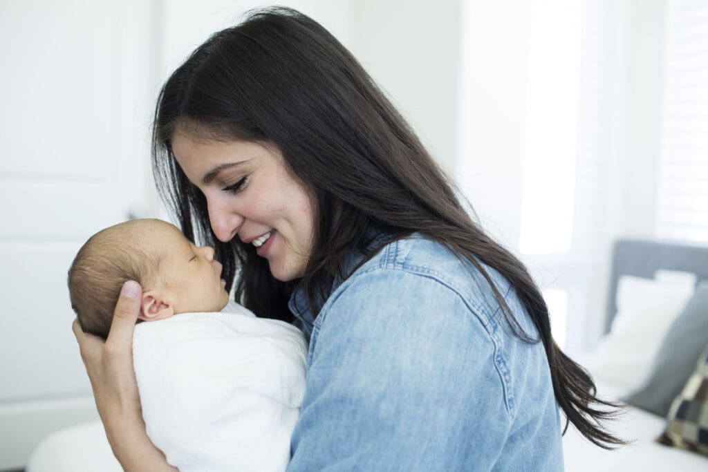 Mom and newborn sitting nose to nose on the edge of the master bed in their brightly lit, modern South Jordan home during an in-home newborn session.
